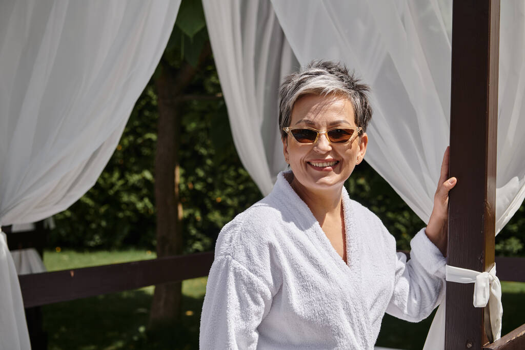 joyful mature woman in sunglasses resting in private pavilion, wellness retreat in luxury resort - Photo, Image