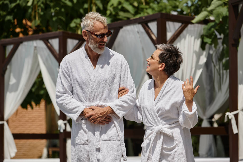 glückliches Paar mittleren Alters in Sonnenbrille und Bademänteln spazieren im Luxus-Resort, Wellness-Rückzugskonzept - Foto, Bild