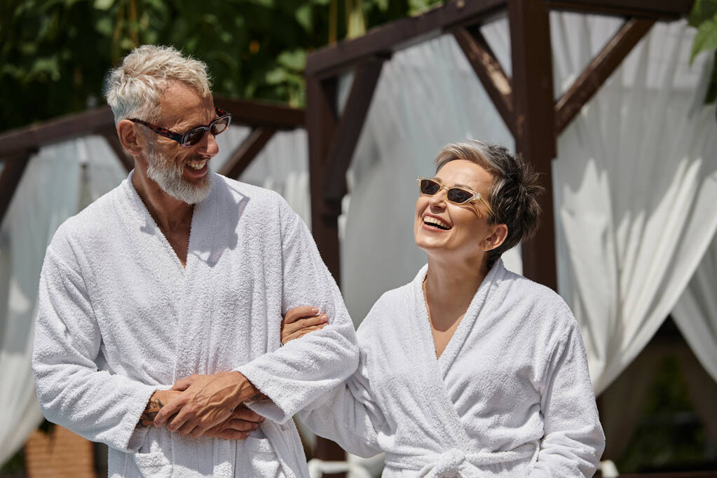 radosna para w średnim wieku w okularach przeciwsłonecznych i szlafrokach spacerujących w luksusowym kurorcie, koncepcja odosobnienia wellness - Zdjęcie, obraz