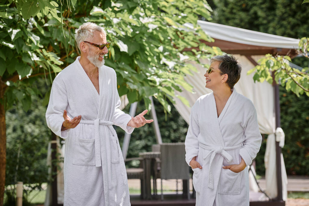 feliz pareja madura en gafas de sol y túnicas charlando en un resort de lujo, concepto de retiro de bienestar - Foto, imagen