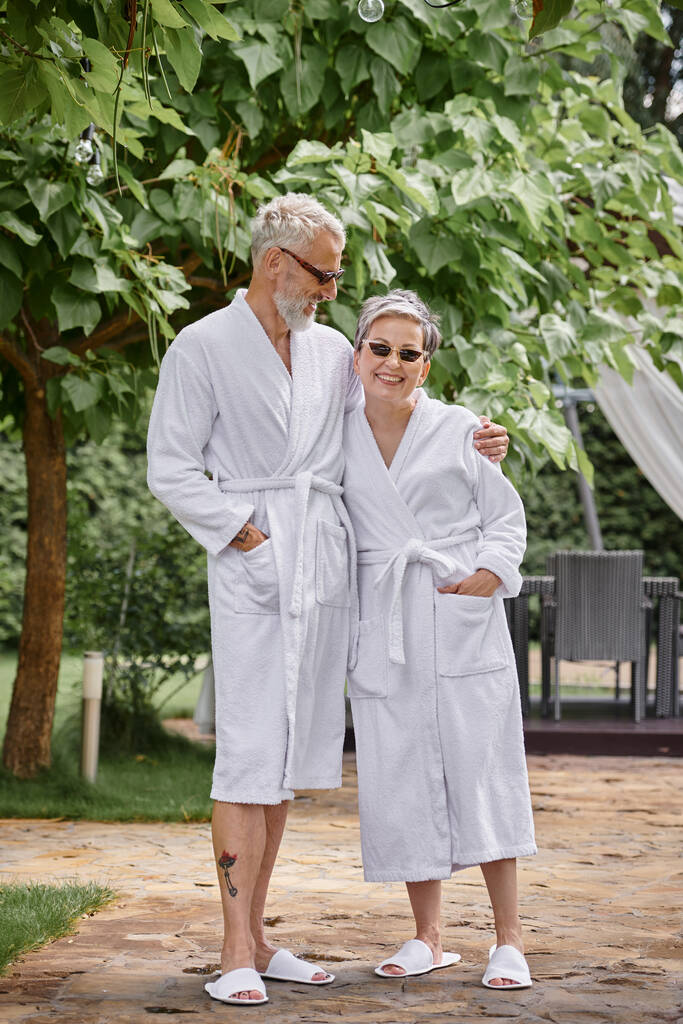 gelukkig volwassen man met tattoo knuffelen vrouw in zonnebril en badjas in de zomer tuin, wellness retraite - Foto, afbeelding