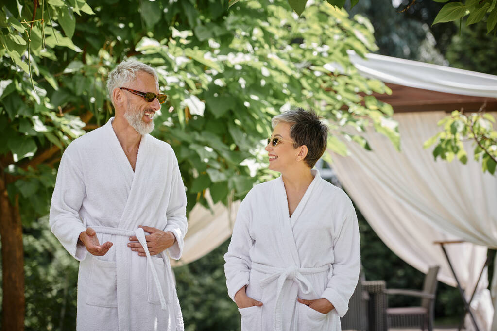 Orta yaşlı bir adam güneş gözlüğü ve bornozlu mutlu eşiyle sohbet ediyor, yaz bahçesi, sağlık inzivası. - Fotoğraf, Görsel