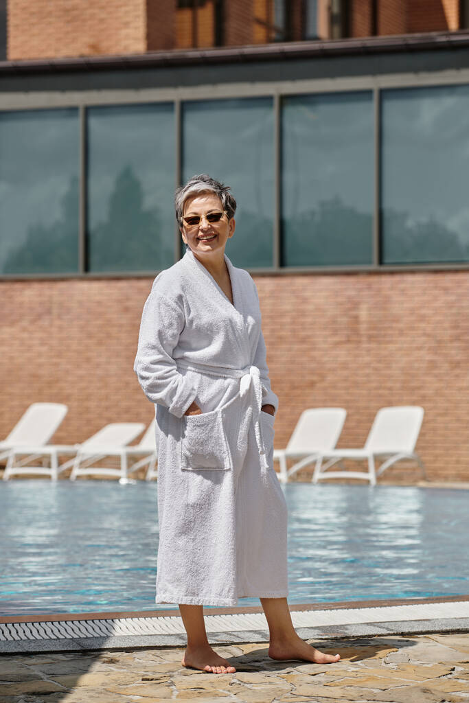 高級リゾートのプールサイドに立っている白いローブとサングラスの幸せな中年女性 - 写真・画像