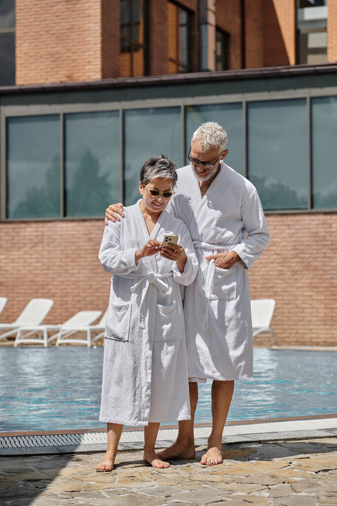 radostná žena středního věku ve slunečních brýlích pomocí smartphonu poblíž manžela u bazénu v luxusním resortu - Fotografie, Obrázek