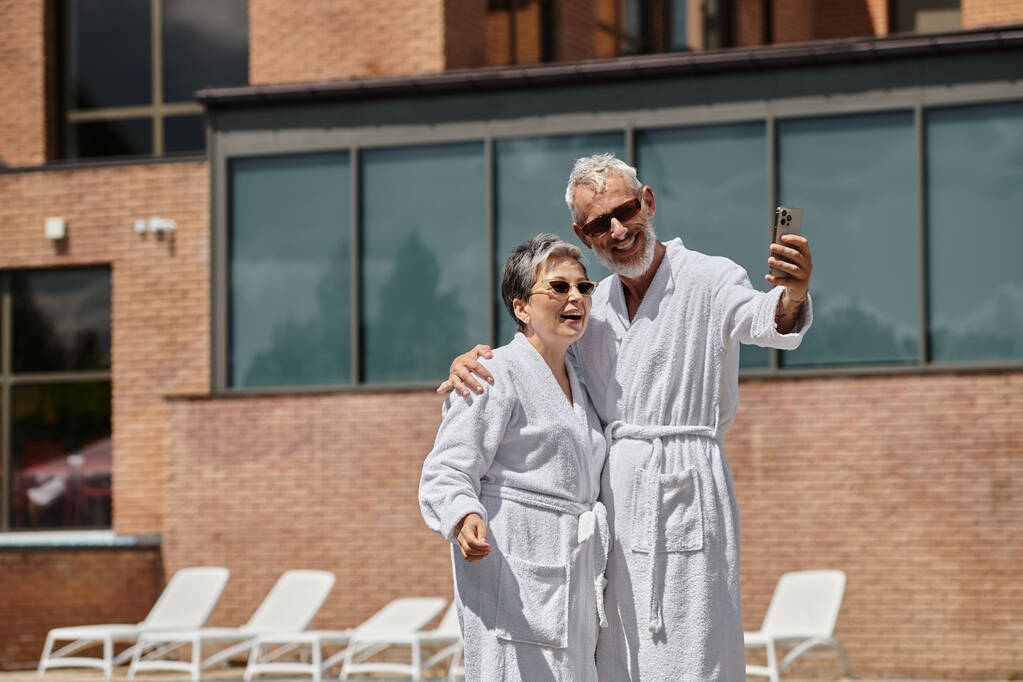 счастливая пара средних лет в халатах делает селфи на смартфоне в роскошном курорте, оздоровительный отдых - Фото, изображение