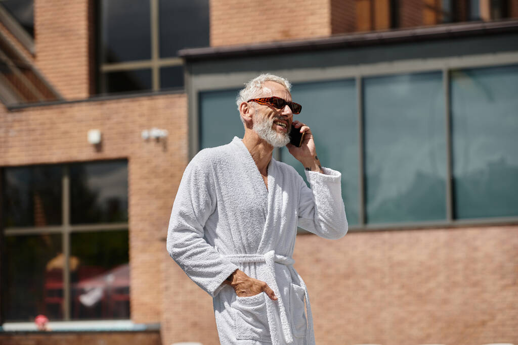 szczęśliwy i wytatuowany mężczyzna w średnim wieku w okularach przeciwsłonecznych i szlafroku rozmawiający na smartfonie w kurorcie, odwrót - Zdjęcie, obraz