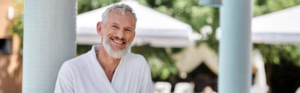 vrolijk volwassen man in witte badjas kijkend naar camera op resort, wellness retraite concept, banner - Foto, afbeelding