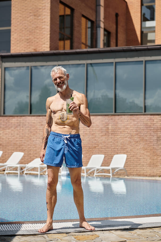 wytatuowany mężczyzna w średnim wieku w szortach uśmiechnięty i trzymający koktajl mojito przy basenie, odpoczynek wellness - Zdjęcie, obraz