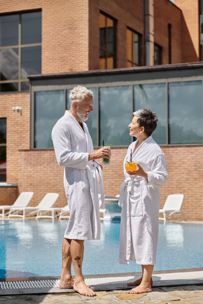 veselý pár středního věku v bílých róbách s koktejly u bazénu, wellness ústup v resortu - Fotografie, Obrázek