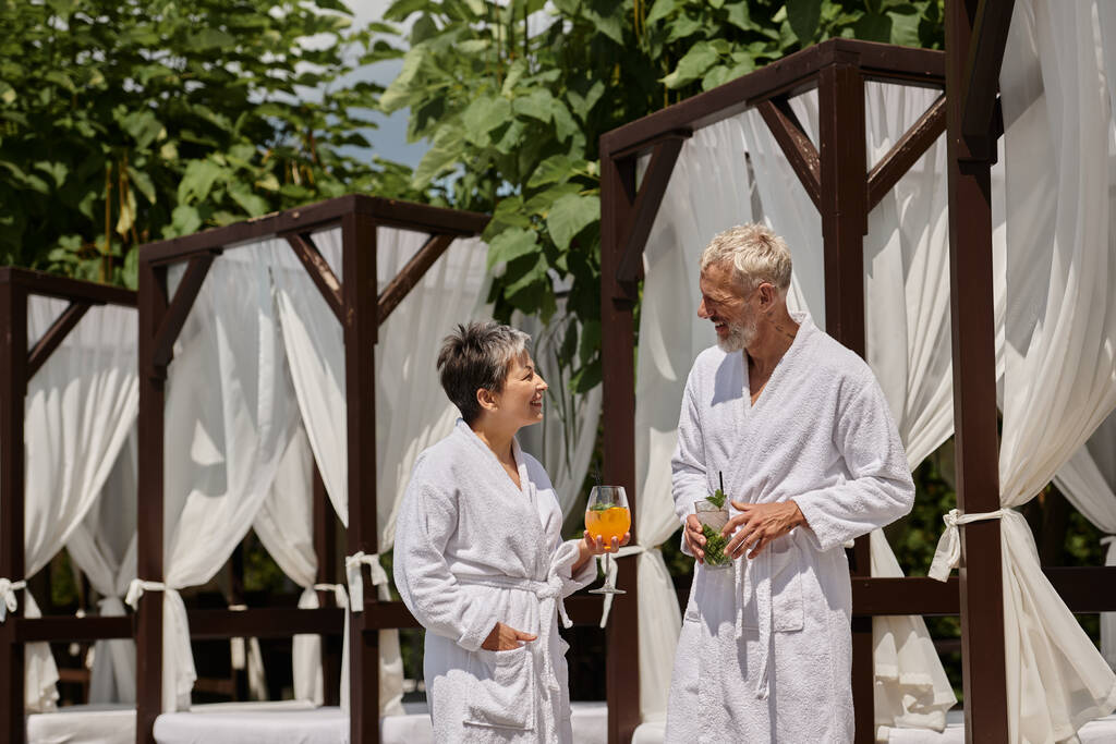 happy mature couple in white robes holding cocktails and talking near pavilion, wellness retreat - Photo, Image