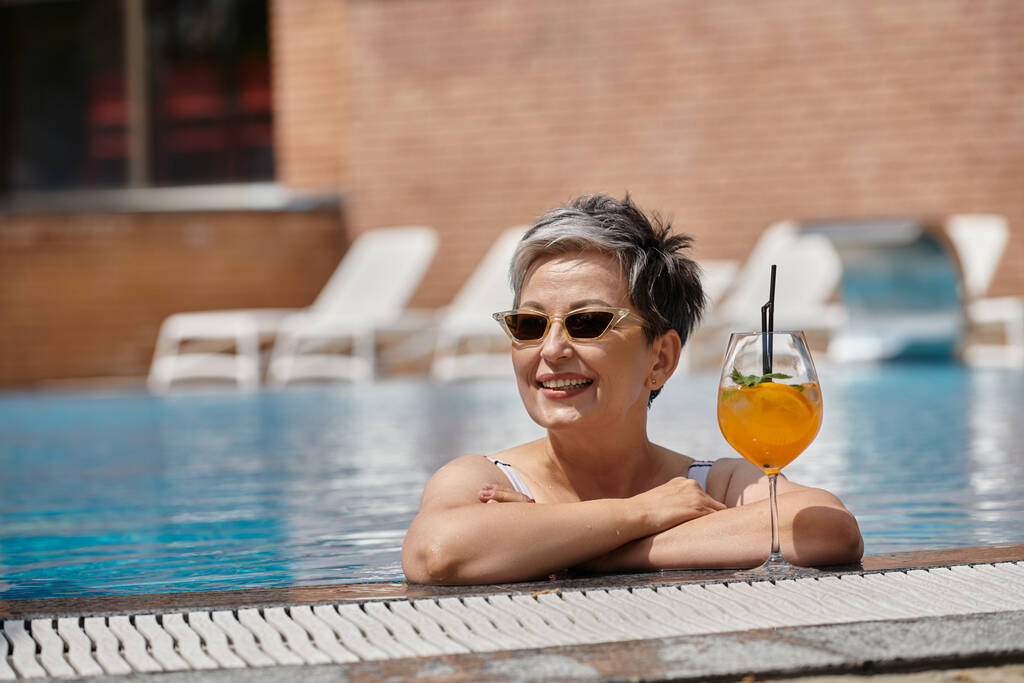 alegre mujer de mediana edad en gafas de sol dentro de la piscina, junto a la piscina con cóctel, retiro - Foto, Imagen