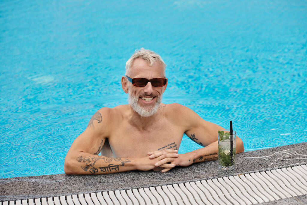 shirtloze en getatoeëerde man van middelbare leeftijd in een zonnebril in het zwembad in de buurt van cocktail, retraite - Foto, afbeelding