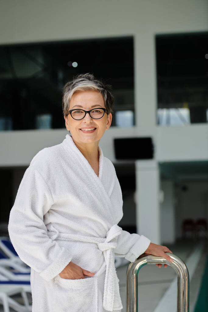 gelukkige vrouw van middelbare leeftijd in witte badjas en bril glimlachen binnen van indoor spa centrum, retraite - Foto, afbeelding