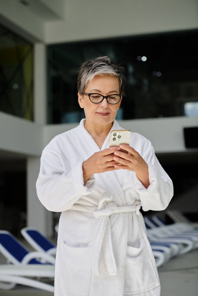 kobieta w średnim wieku w białym szlafroku i okularach za pomocą smartfona wewnątrz krytego centrum spa, rekolekcje - Zdjęcie, obraz