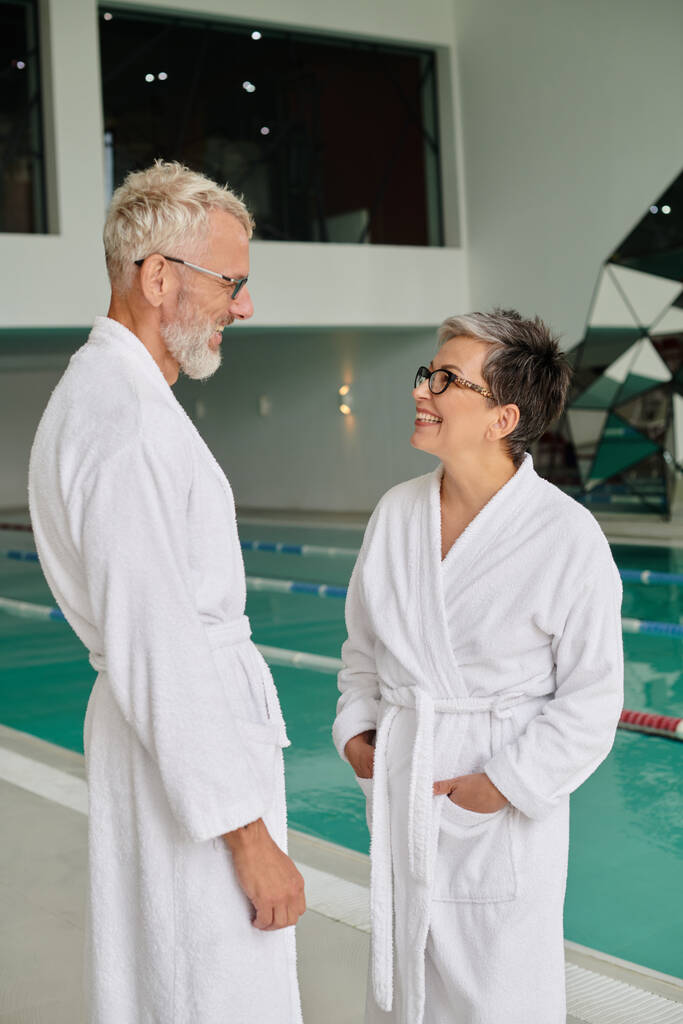 vrolijk stel van middelbare leeftijd in bril en witte gewaden staan in de buurt van een overdekt zwembad in spa centrum - Foto, afbeelding