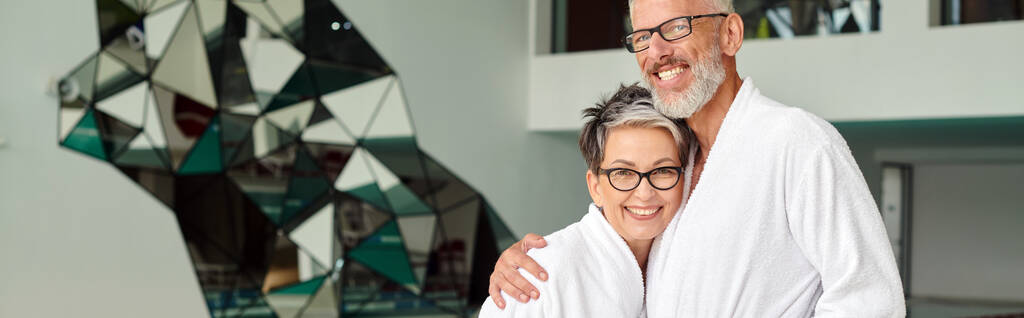 Gözlüklü ve beyaz cüppeli mutlu orta yaşlı adam spa merkezinde karısını kucaklıyor, sağIık inzivası, pankart - Fotoğraf, Görsel