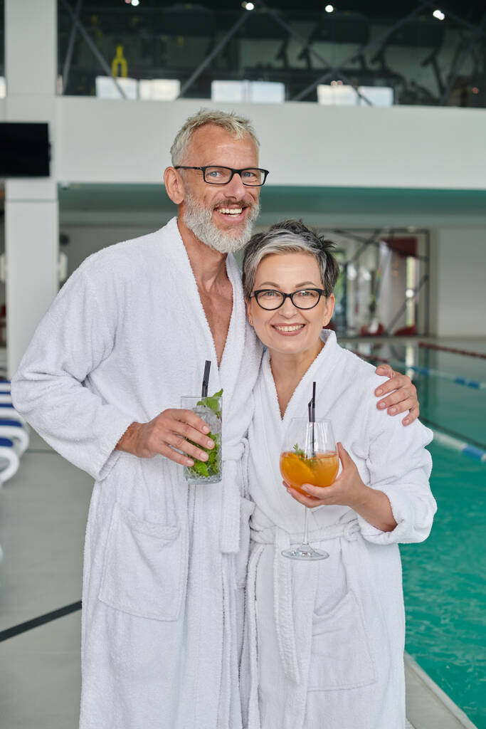 glückliches Paar mittleren Alters in weißen Roben mit Gläsern mit Cocktails im Wellness-Center in der Nähe des Pools - Foto, Bild