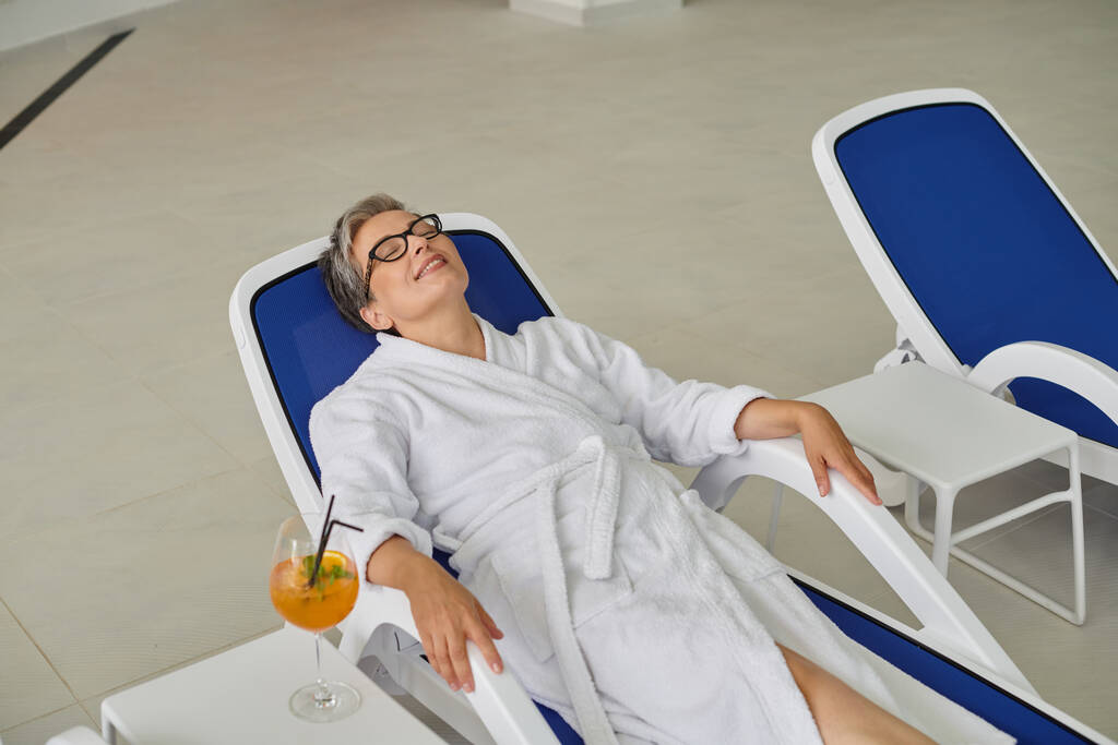 retreat concept, pleased mature woman in white robe resting on lounger near cocktail in spa center - Photo, Image