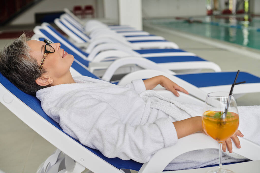 conceito retiro spa, mulher madura satisfeito em roupão branco e óculos descansando na espreguiçadeira perto de coquetel - Foto, Imagem