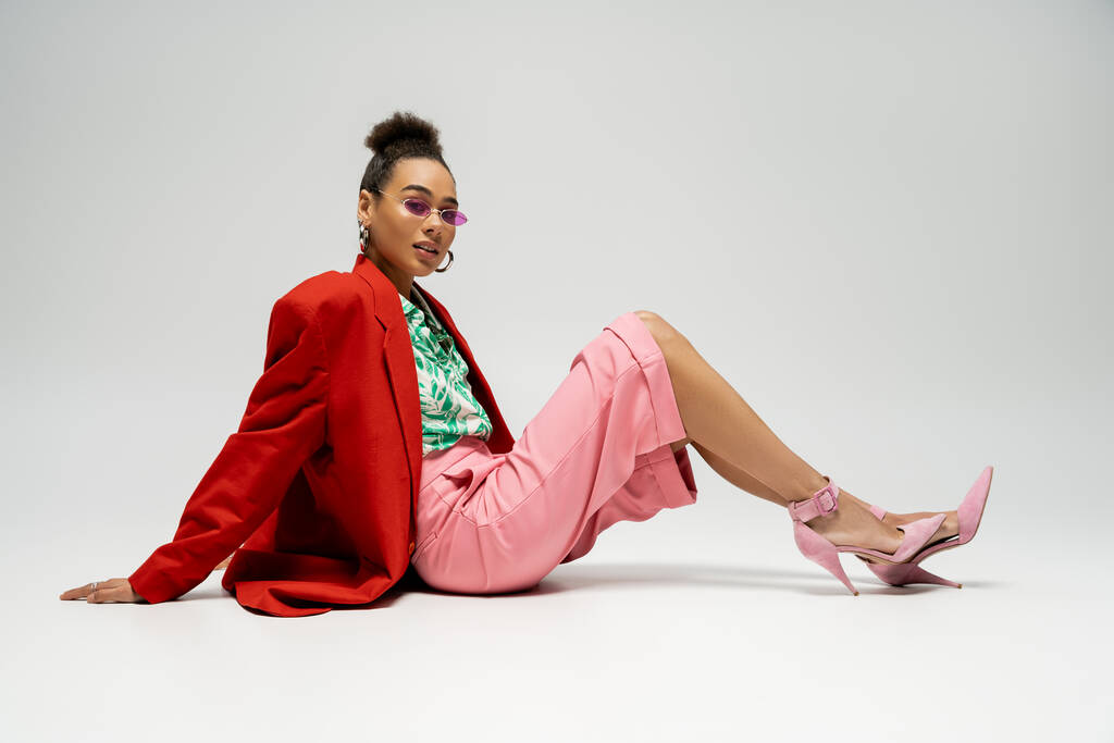 уверенная африканская американка в стильной одежде и розовых солнцезащитных очках сидит на сером фоне - Фото, изображение