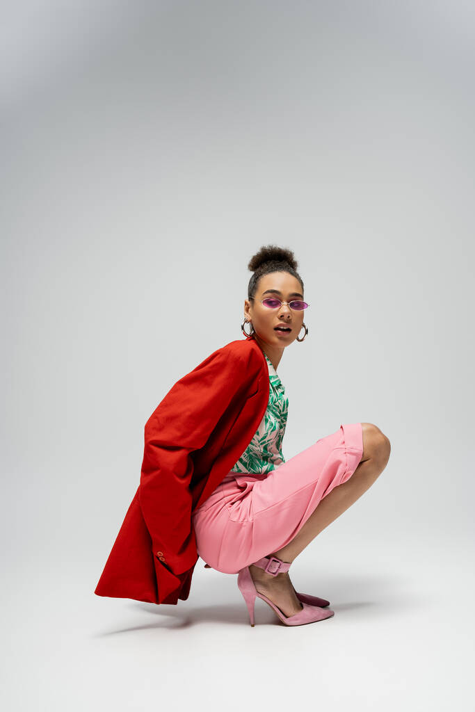 mladý africký americký model ve stylovém oblečení a růžové sluneční brýle sedí na šedém pozadí - Fotografie, Obrázek
