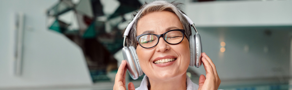 χαρούμενη μεσήλικη γυναίκα με γυαλιά που ακούει μουσική σε ασύρματα ακουστικά στο κέντρο σπα, banner - Φωτογραφία, εικόνα