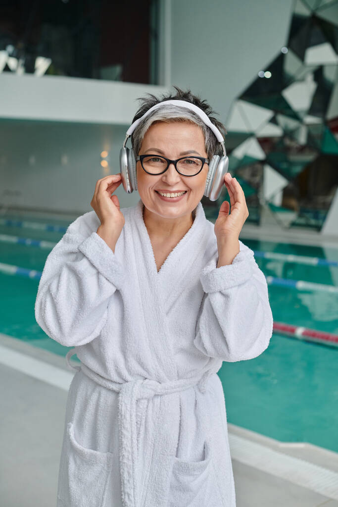 happy middle aged woman in glasses listening music in wireless headphones in spa center, retreat - Photo, Image