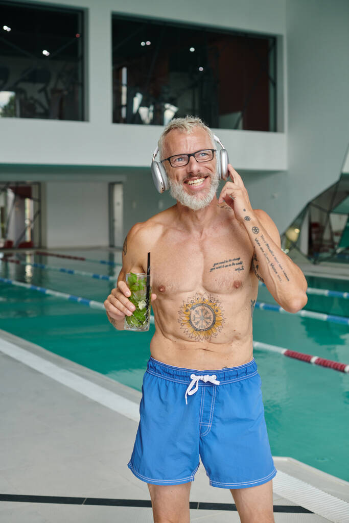 retiro, hombre de mediana edad tatuado escuchando música en auriculares y celebrando cóctel en el centro de spa - Foto, Imagen