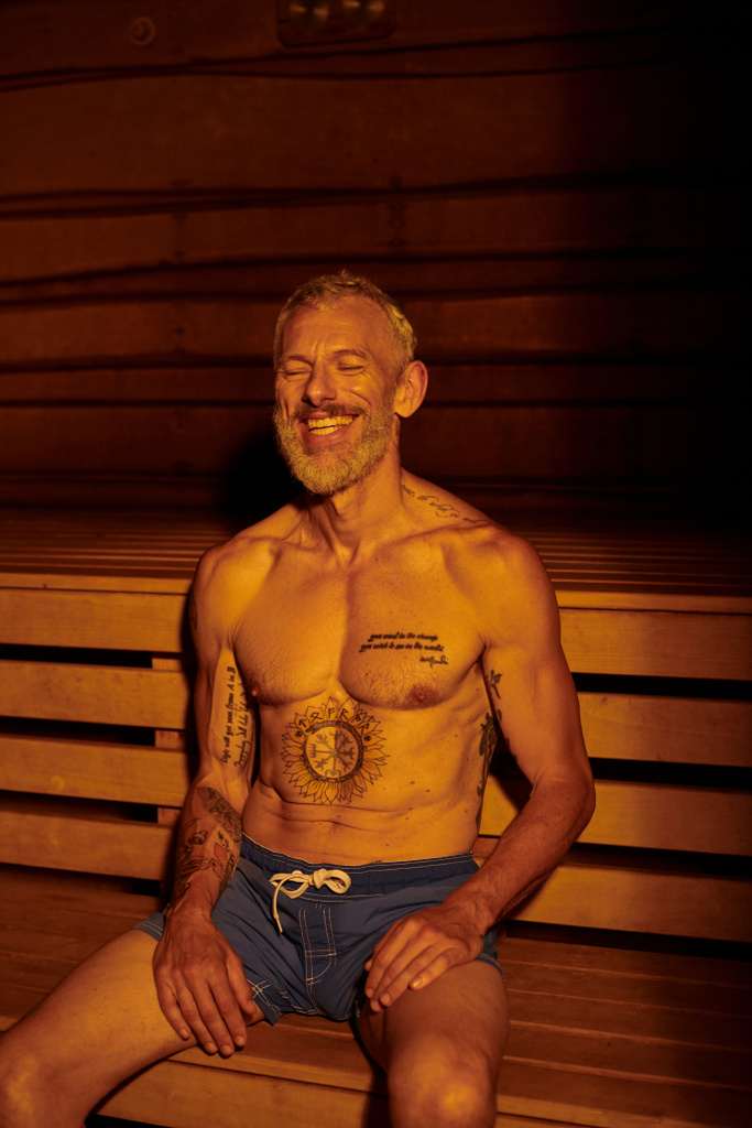 homme d'âge moyen heureux et torse nu avec tatouages assis dans le sauna, concept de retraite bien-être - Photo, image