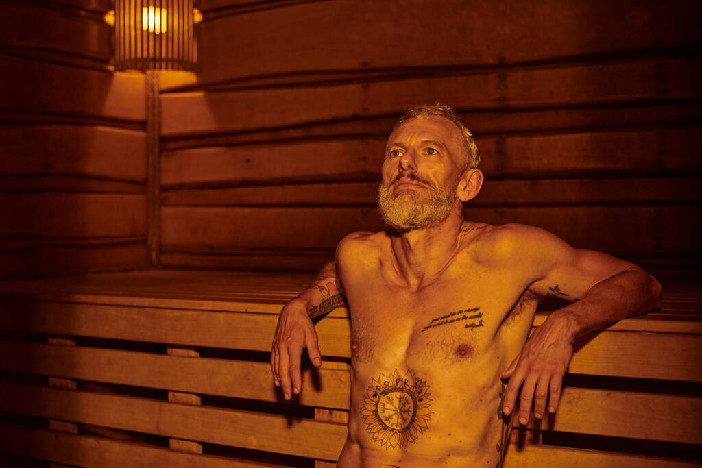 hombre de mediana edad pensativo y sin camisa con tatuajes sentado en la sauna, concepto de retiro de bienestar - Foto, Imagen