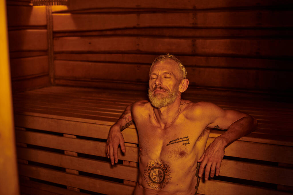 hombre de mediana edad tranquilo, relajado y sin camisa con tatuajes sentado en la sauna, retiro de bienestar - Foto, Imagen