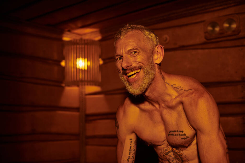 hombre de mediana edad alegre y sin camisa con tatuajes sentado en la sauna, concepto de retiro de bienestar - Foto, Imagen