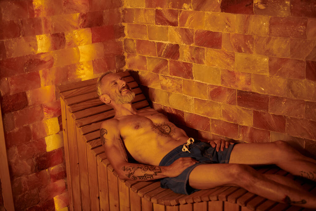 vidám és félmeztelen középkorú férfi tetoválásokkal a szaunában, wellness elvonulás koncepció - Fotó, kép