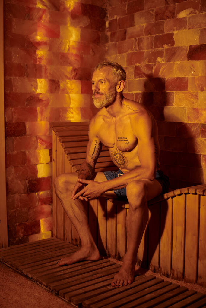 uvolněný a bez trička muž středního věku s tetováním sedí na lavičce v sauně, wellness koncept - Fotografie, Obrázek
