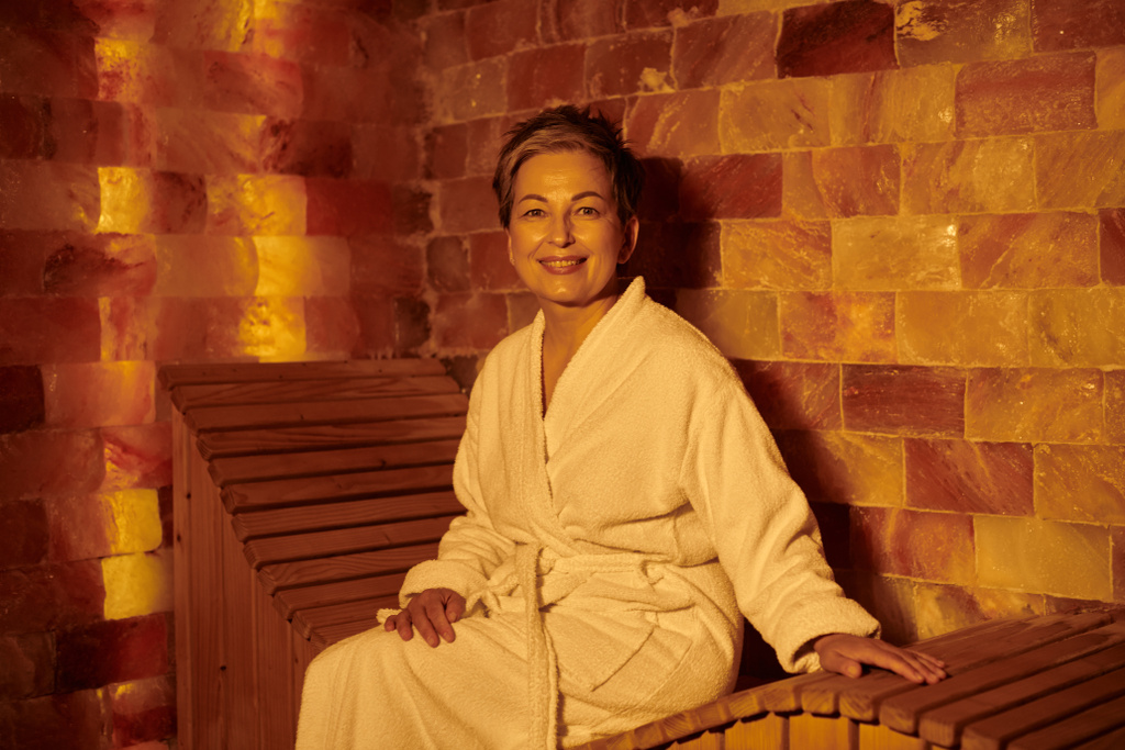 feliz mujer de mediana edad en bata blanca sentado en el banco en la sauna, spa concepto de bienestar, retiro - Foto, Imagen