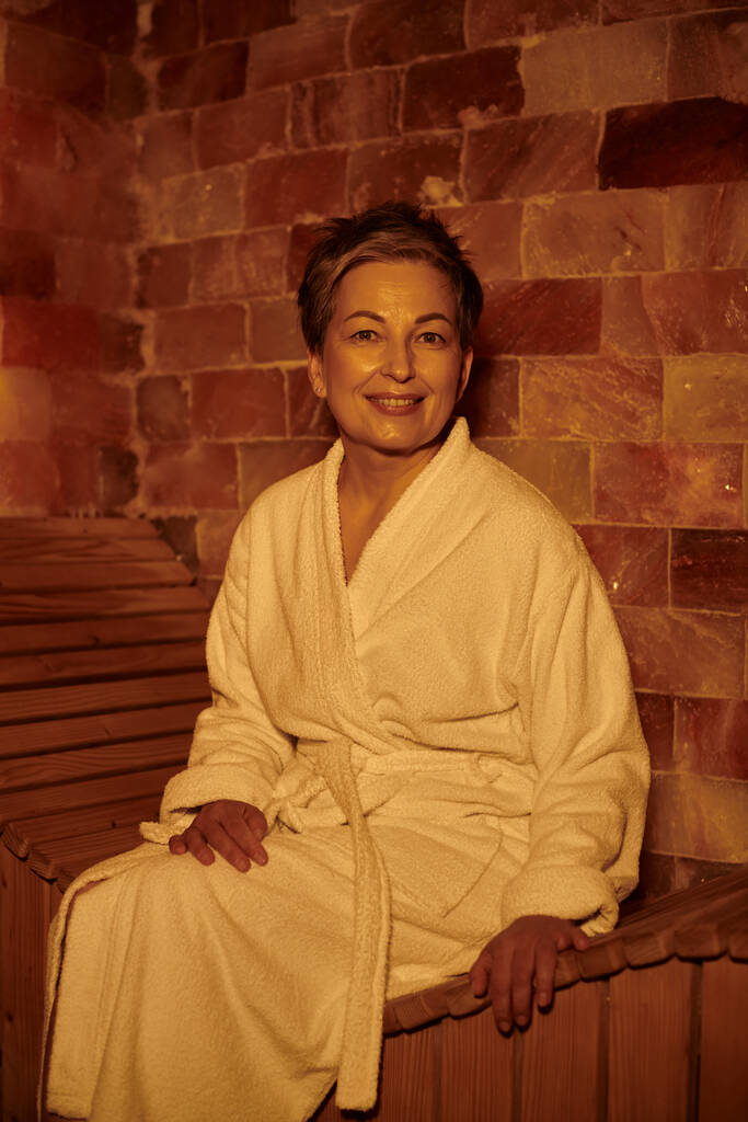 vidám középkorú nő fehér köntösben ül padon szaunában, spa wellness koncepció, visszavonulás - Fotó, kép