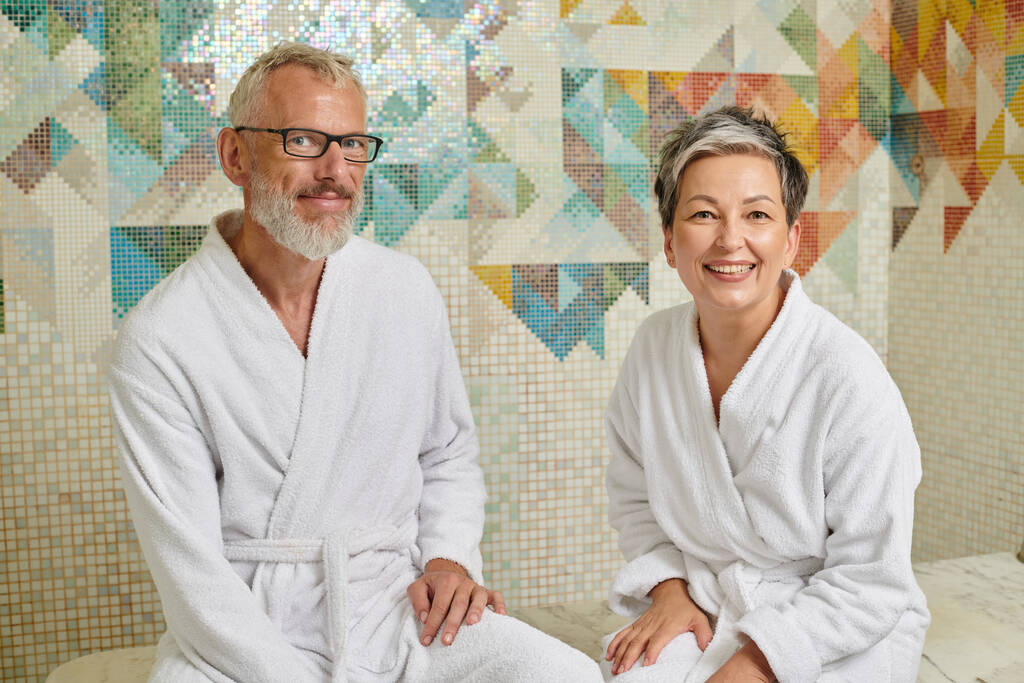 alegre pareja de mediana edad en túnicas blancas sentado en la sauna, spa y el concepto de bienestar, hammam - Foto, Imagen