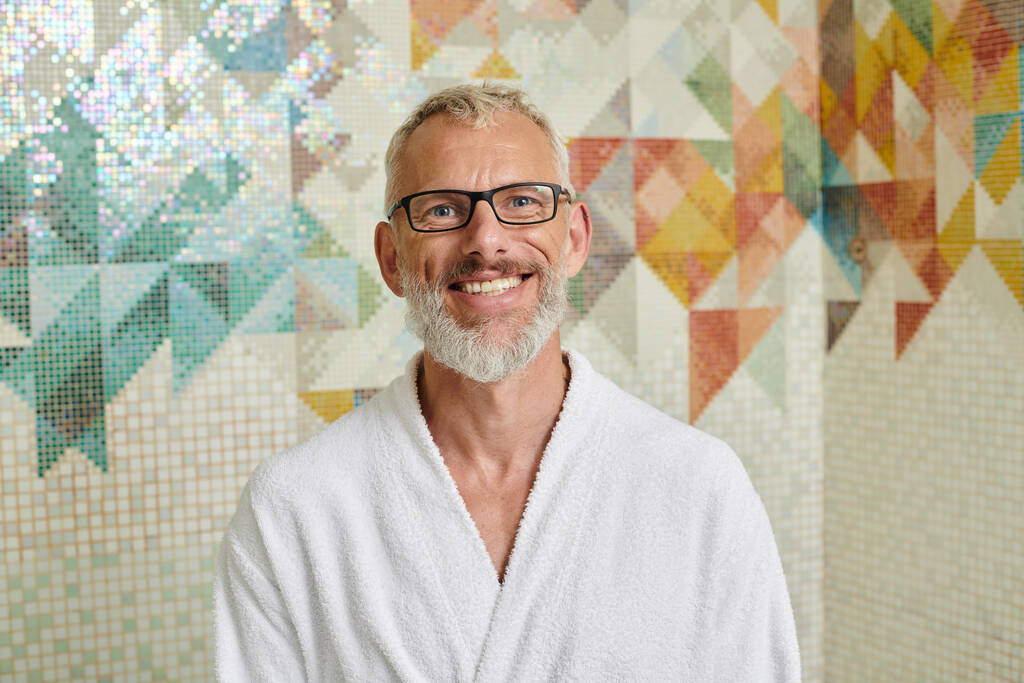 joyful middle aged man in white robe sitting in sauna, spa and wellness concept, marble hammam - Photo, Image