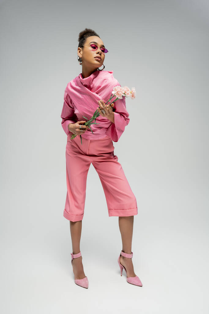 εκφραστικό αφρικανικό μοντέλο μόδας σε ροζ ενδυμασία και τακούνια που ποζάρουν με λουλούδια, ολόσωμο - Φωτογραφία, εικόνα