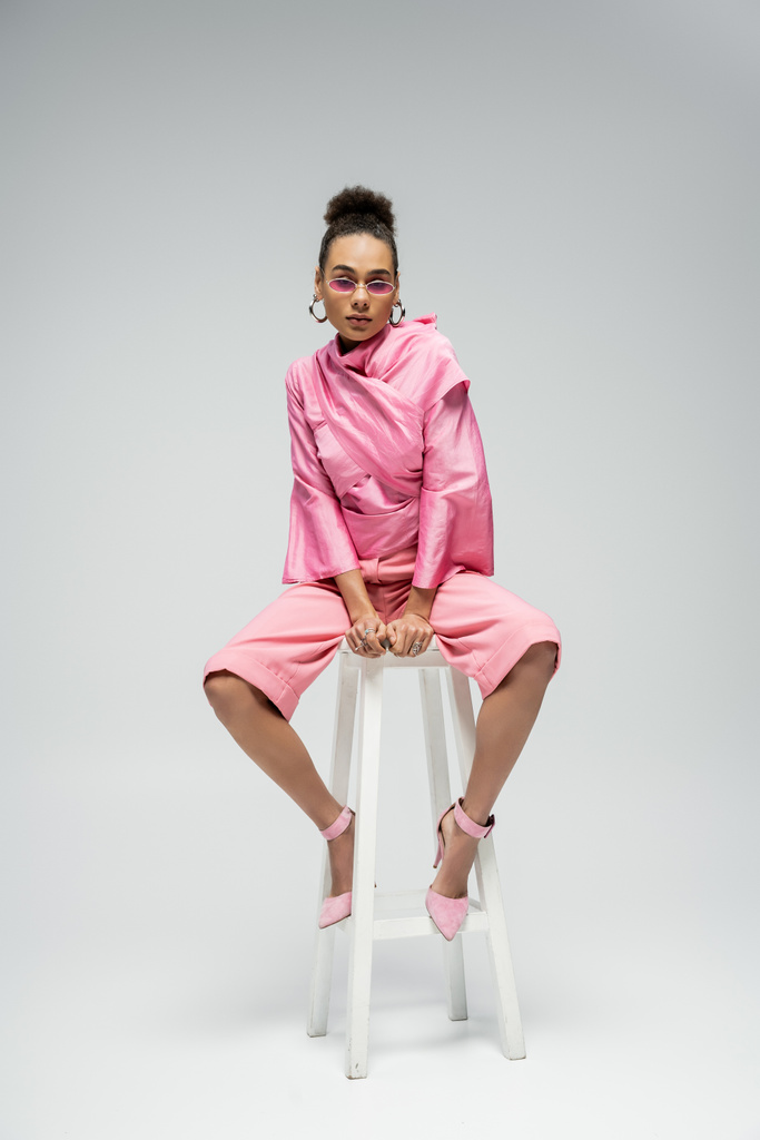 Afrikai amerikai divat modell rózsaszín ruhában és napszemüveg ül a széken, és pózol a szürke - Fotó, kép