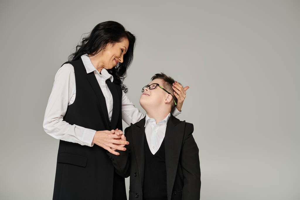 žena v obchodním oblečení a syn s Downovým syndromem ve školní uniformě při pohledu na sebe na šedé - Fotografie, Obrázek