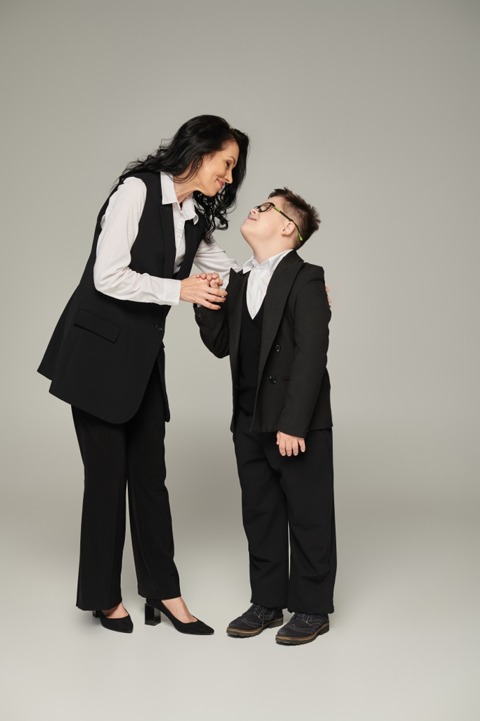 zakenvrouw en zoon met down syndroom in schooluniform hand in hand en glimlach op grijs - Foto, afbeelding