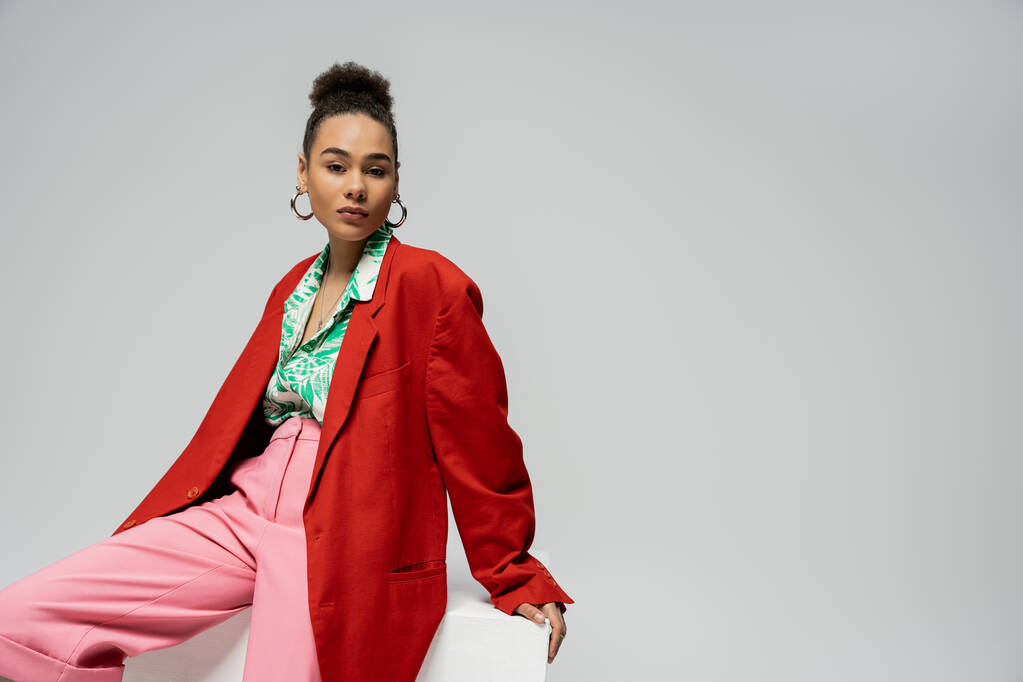 stylish african american model in trendy and vibrant outfit sitting on cube on grey backdrop - Photo, Image