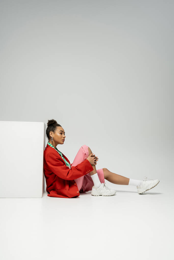 вдумчивая африканская американка в современной и яркой одежде сидит рядом с кубом на сером фоне - Фото, изображение