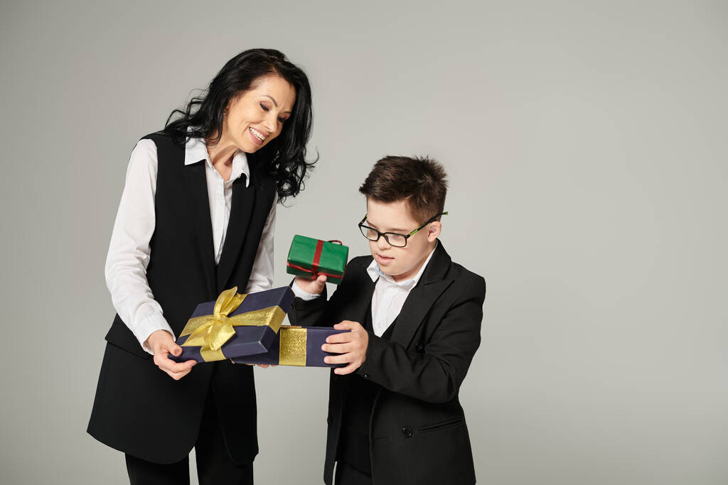 tiener schooljongen met naar beneden syndroom opening geschenk doos in de buurt gelukkig moeder in formele slijtage op grijs - Foto, afbeelding