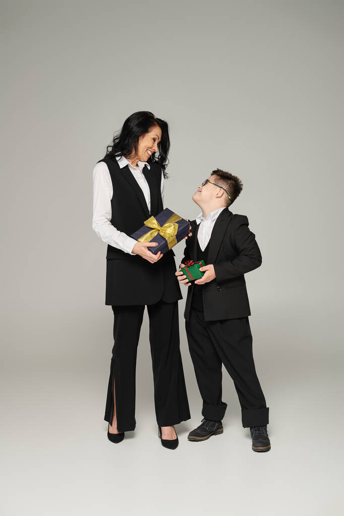 bizneswoman i szczęśliwy syn z zespołem Downa w mundurze szkolnym trzymając pudełka z prezentami na szarości - Zdjęcie, obraz