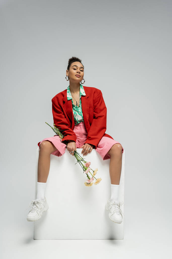 african american woman in stylish red blazer holding flowers and sitting on cube on grey backdrop - Photo, Image