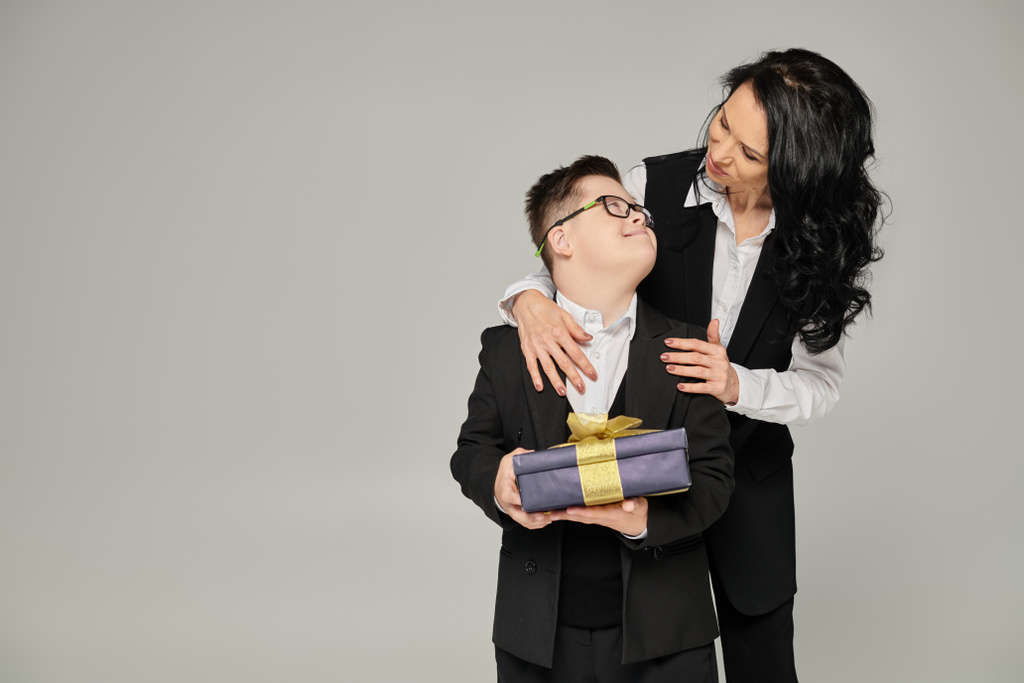 glücklich gekleidete Frau umarmt Sohn mit Down-Syndrom und hält Geschenkschachtel auf grauem, einzigartigem Familienbild - Foto, Bild
