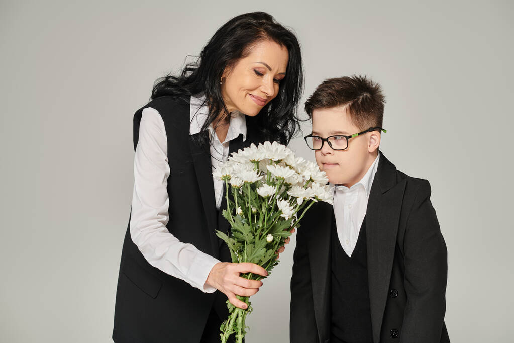 радостная женщина держит цветы рядом с сыном с синдромом Дауна в школьной форме и очки на сером - Фото, изображение