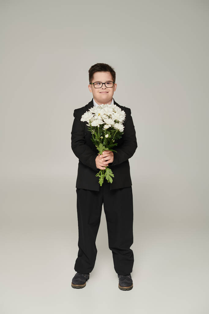 ragazzo con sindrome di Down in uniforme scolastica e occhiali che reggono fiori e sorridono sul grigio - Foto, immagini